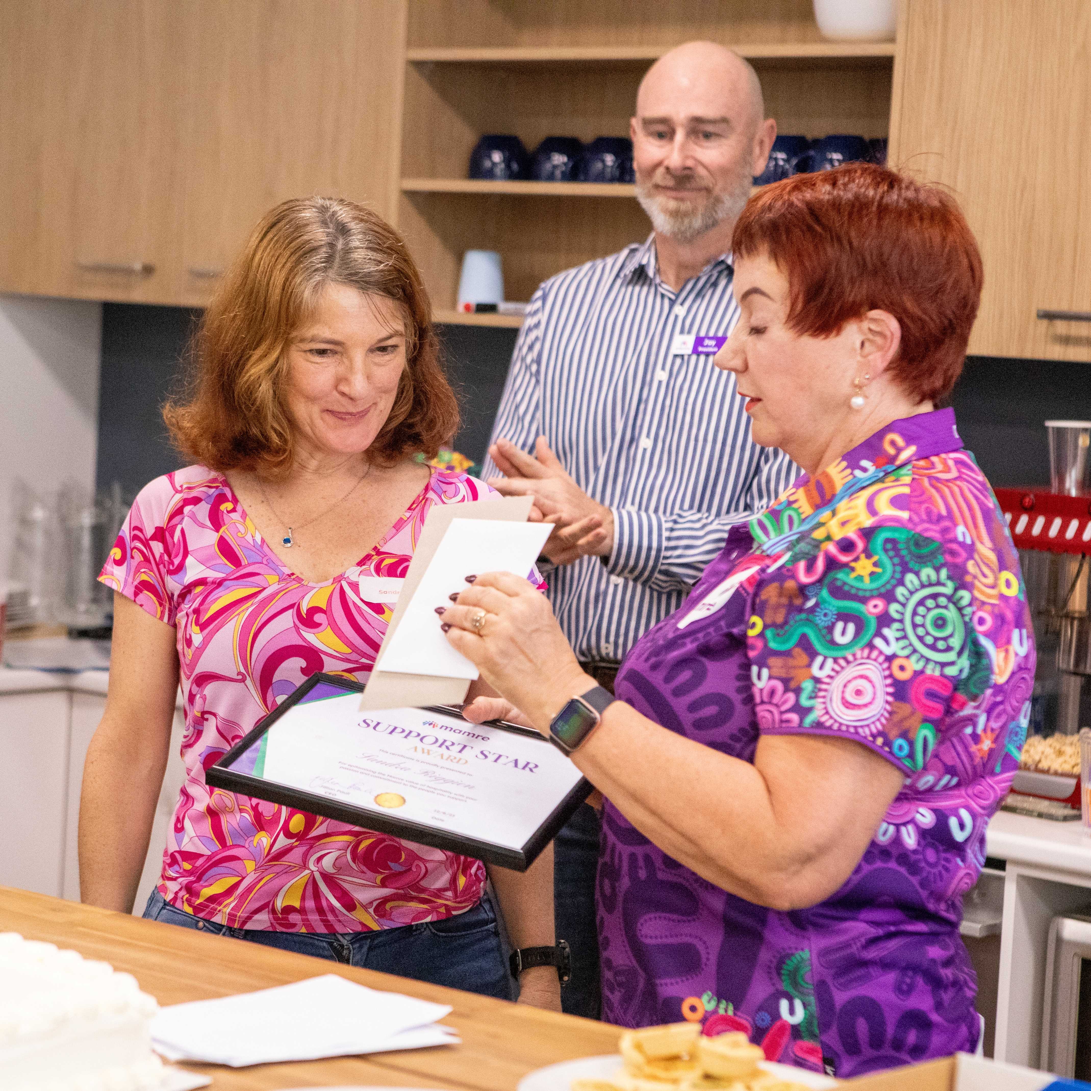 Support Worker Sandy receives a certificate and gift for the Support Star Award from Mamre CEO Jillian Paull while Mamre Board President Jay Emmerton watches in the background.