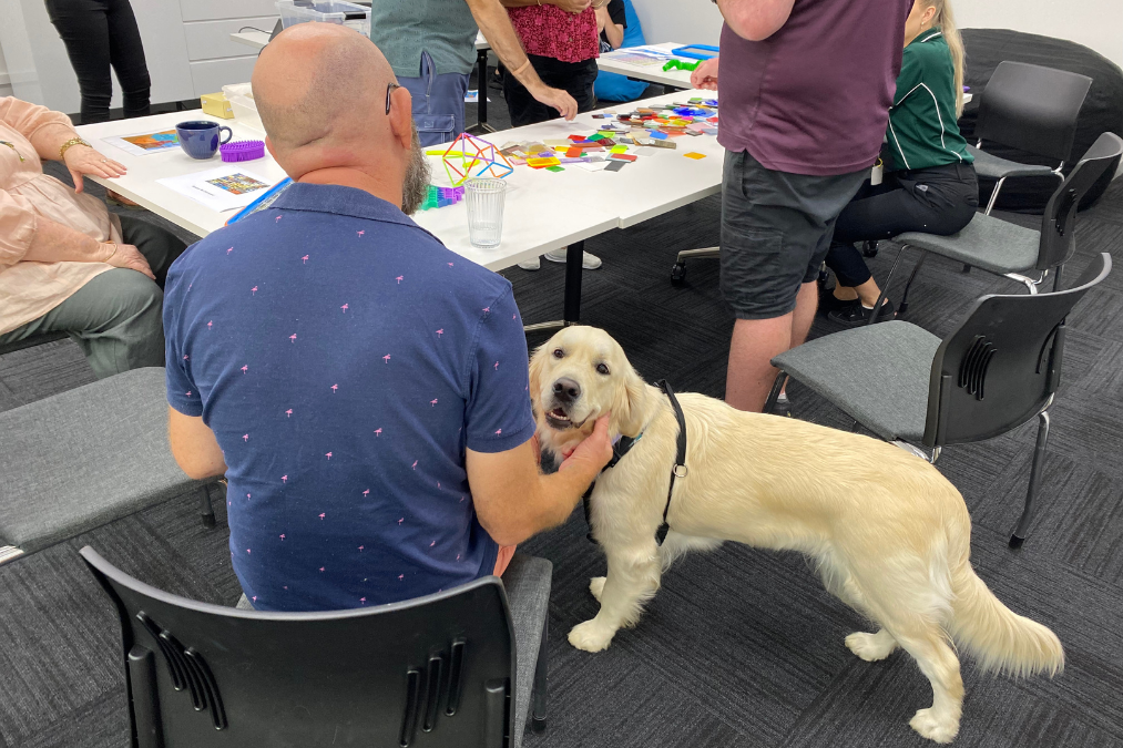 A man pets an Allied Health therapy dog while sitting at a table of arts and crafts with other people.